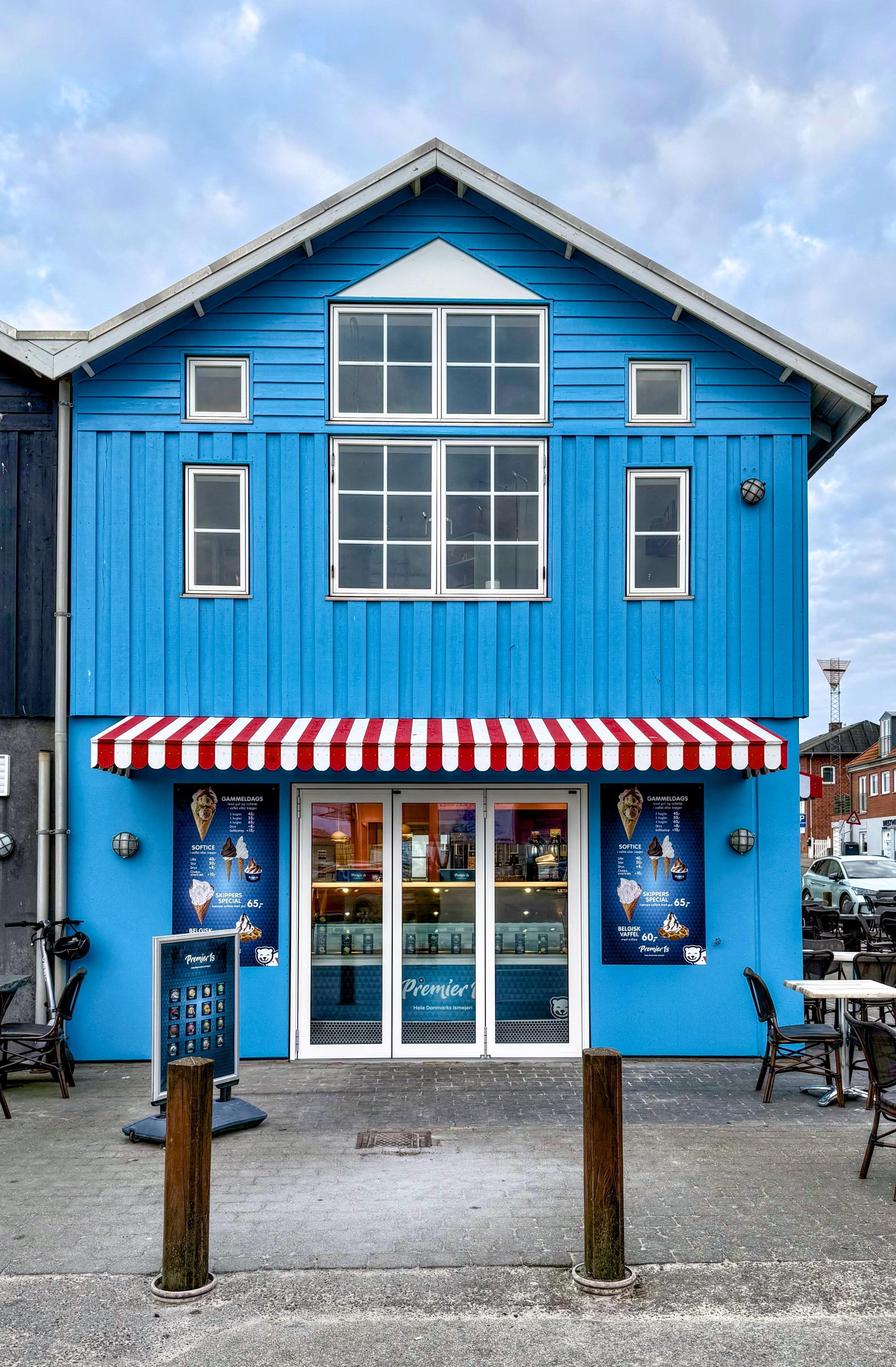Bright blue two-story building with a triangular roof and white frame windows. The ground floor features a glass door flanked by two large windows, and an awning with red and white stripes above them. Large pictures of ice cream and a digital menu are visible on the facade near the entrance. Outdoor seating is present on the left side, and two wooden posts are in the foreground.