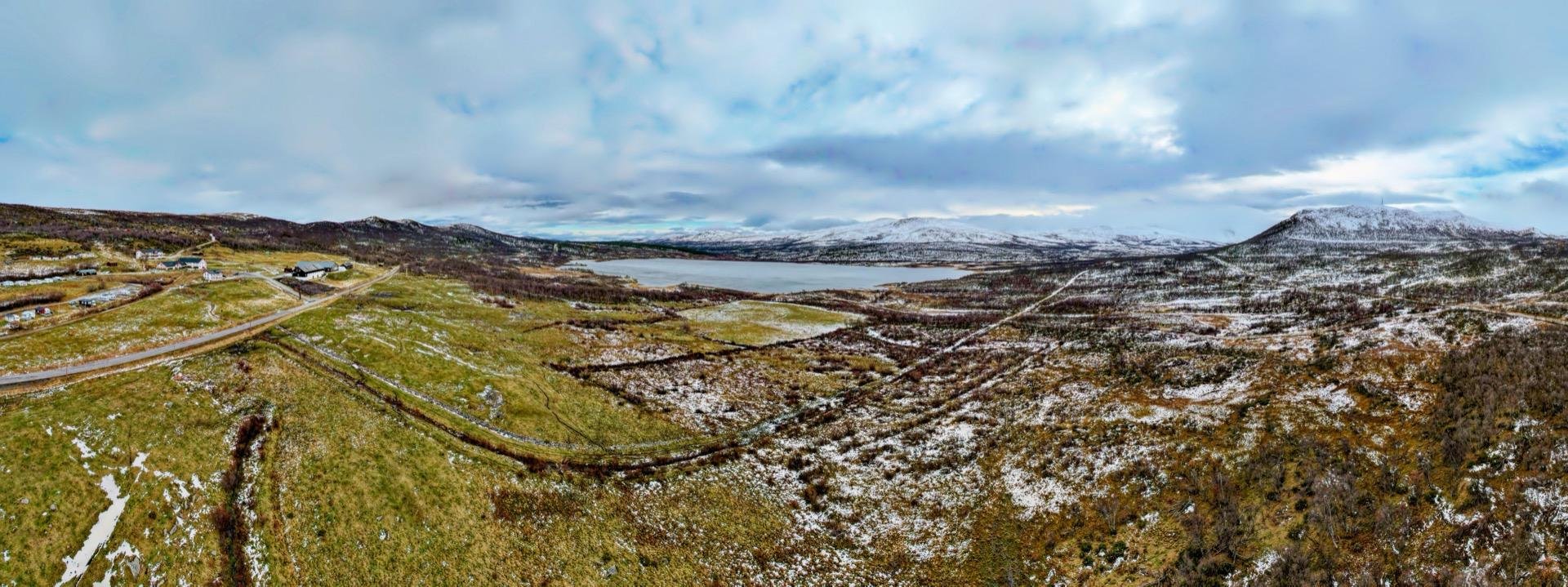 Panoramic view of a snowy landscape featuring undulating hills and a sparsely vegetated valley. A road weaves through the terrain, adjacent to a large body of water bordered by mountains. Overcast skies suggest cold weather conditions, enhancing the muted tones of the natural setting.