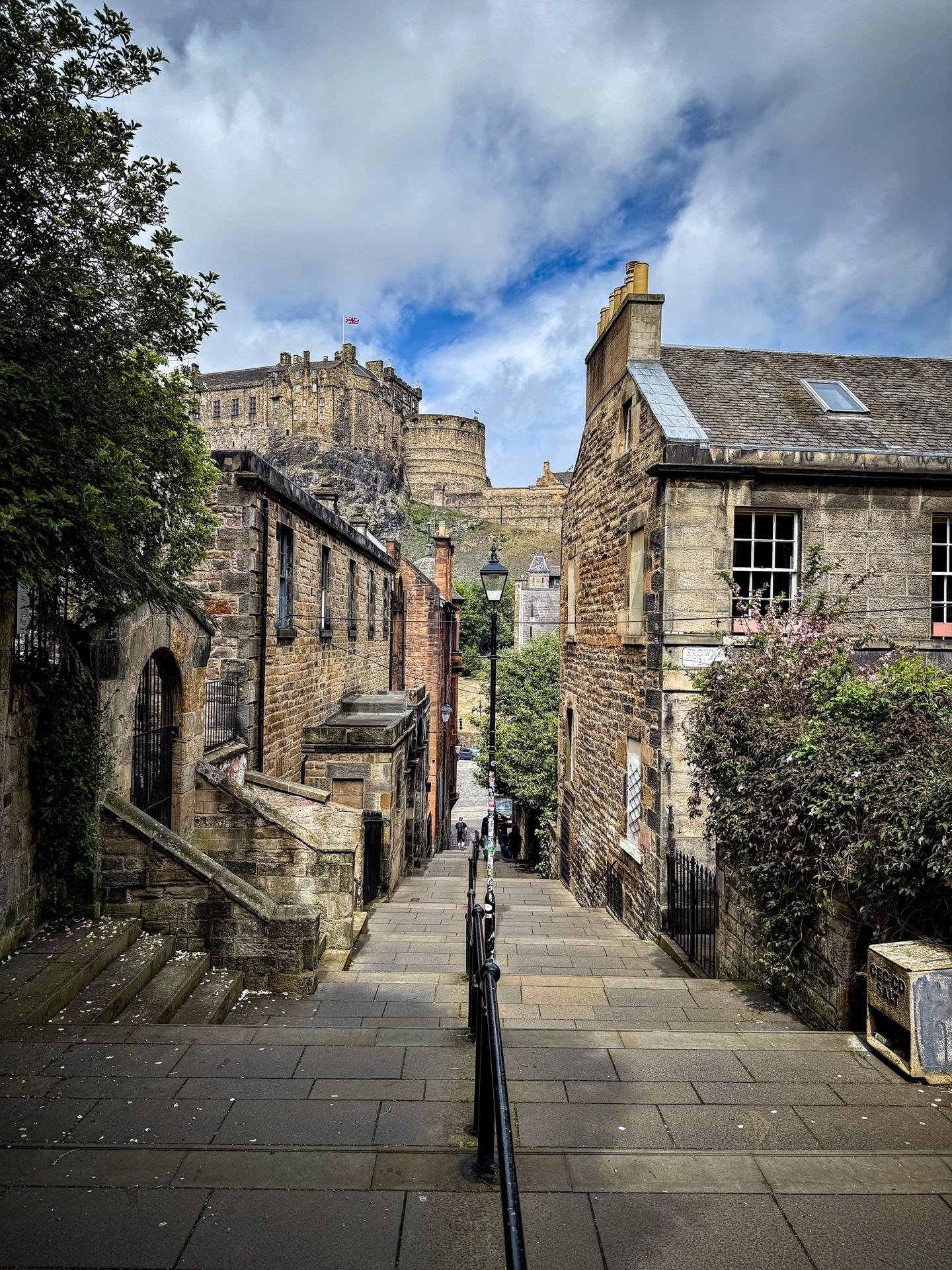 Narrow cobblestone street leading towards a large, ancient castle perched on a rocky hill, flanked by traditional stone buildings with slate roofs. A metal railing runs down the left side of the street. The sky is partly cloudy, and there are lush green trees visible on the edges of the frame.