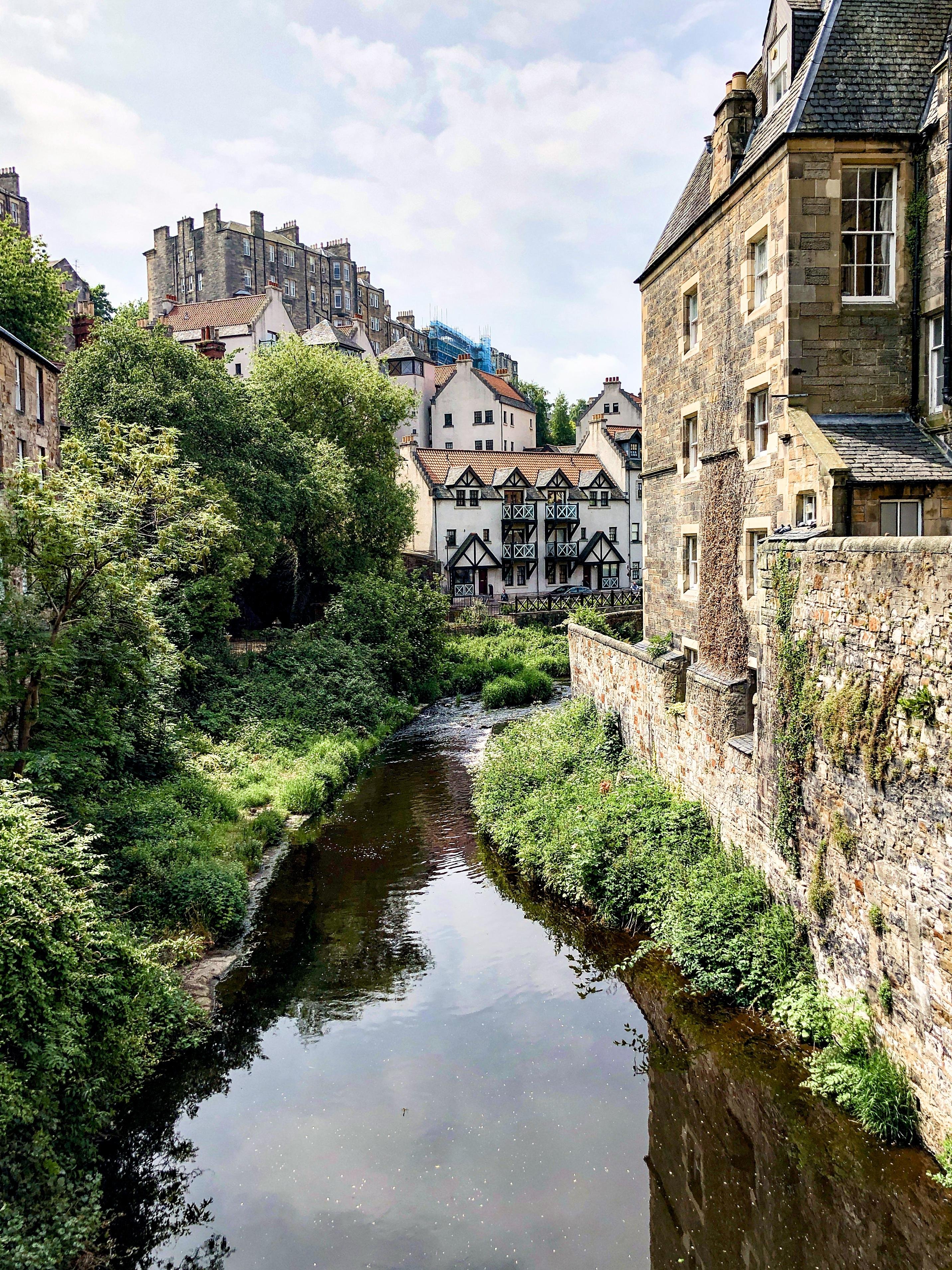 Narrow river flanked by lush greenery and old stone buildings, with a mix of architectural styles including traditional timber framing. The scene is set in an urban area, apparent from the tall, densely packed buildings in the background, some partially obscured by trees.