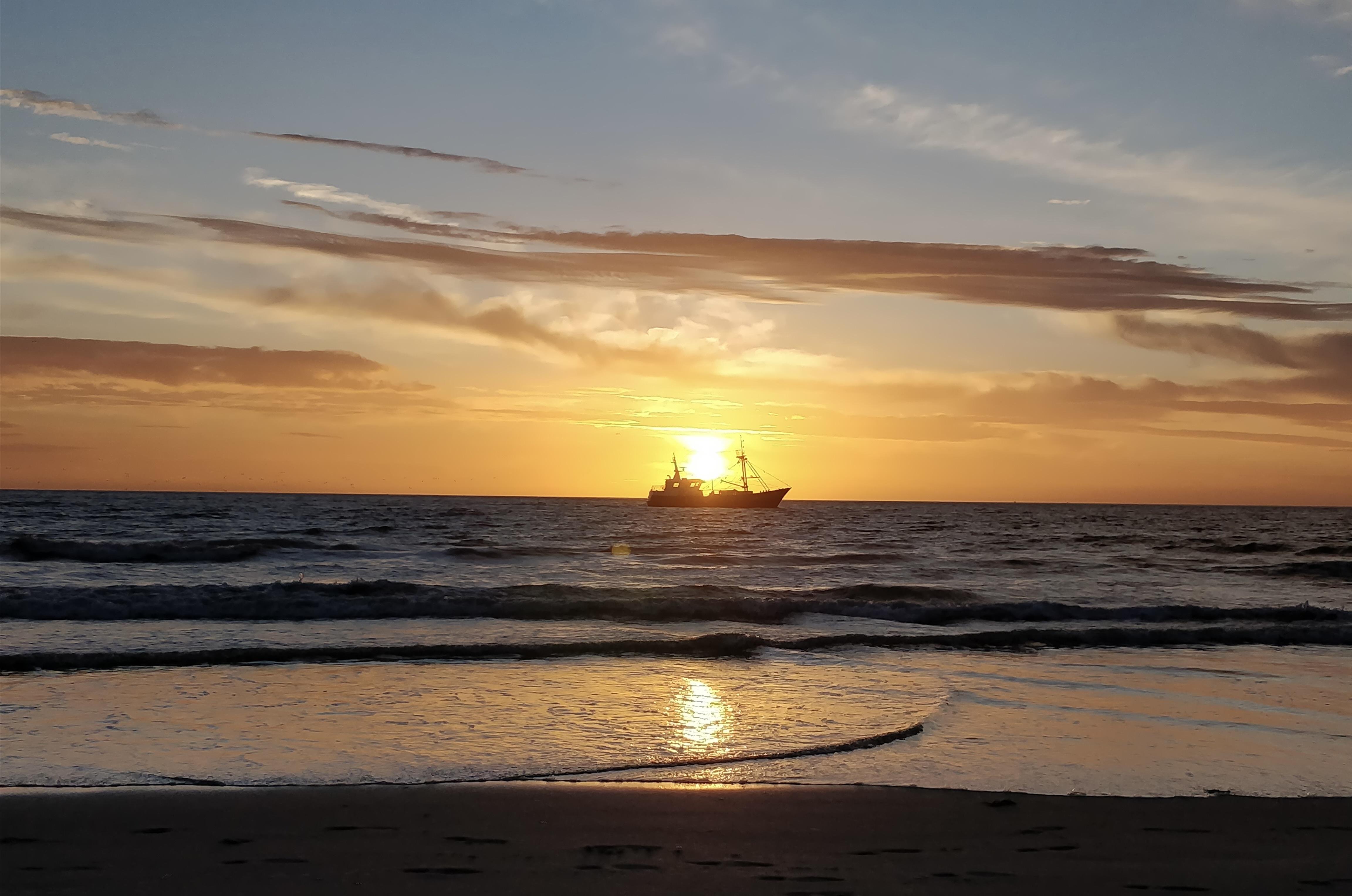 Sunset over the ocean with two silhouetted ships on the horizon, the sun positioned directly above the closer ship. The sky displays a gradient of orange and blue, with scattered clouds. Waves gently approach a sandy beach in the foreground.