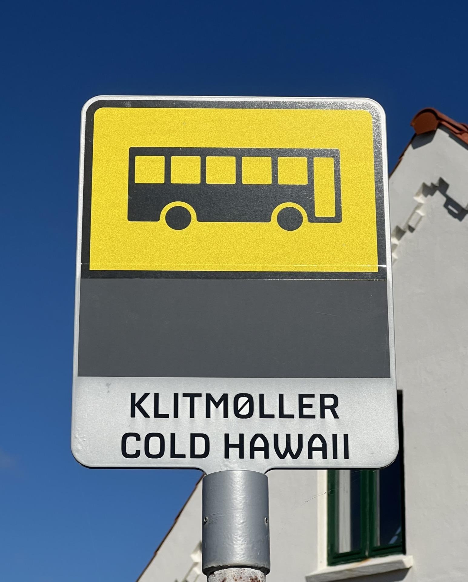Bus stop sign featuring a yellow and black graphic of a bus on a blue sky background, with a text label reading "Klitmøller Cold Hawaii" on a gray background below.