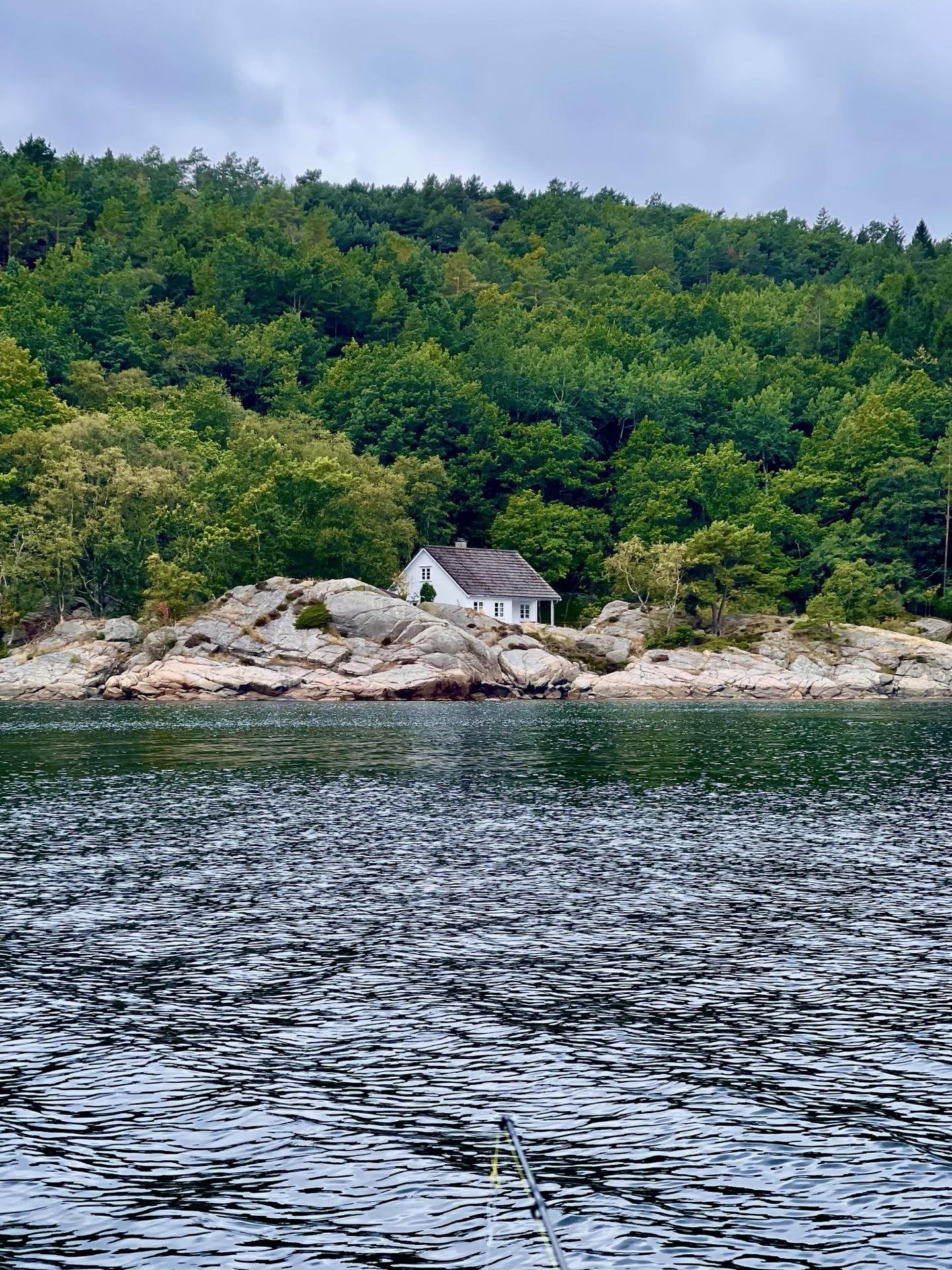 A small white house with a steeply pitched roof is nestled among large rocks on a shoreline, with a dense forest of green trees on the hill behind it. The foreground shows rippling water, reflecting hints of grey under a cloudy sky. A part of a fishing rod is visible at the bottom of the image, extending into the water.