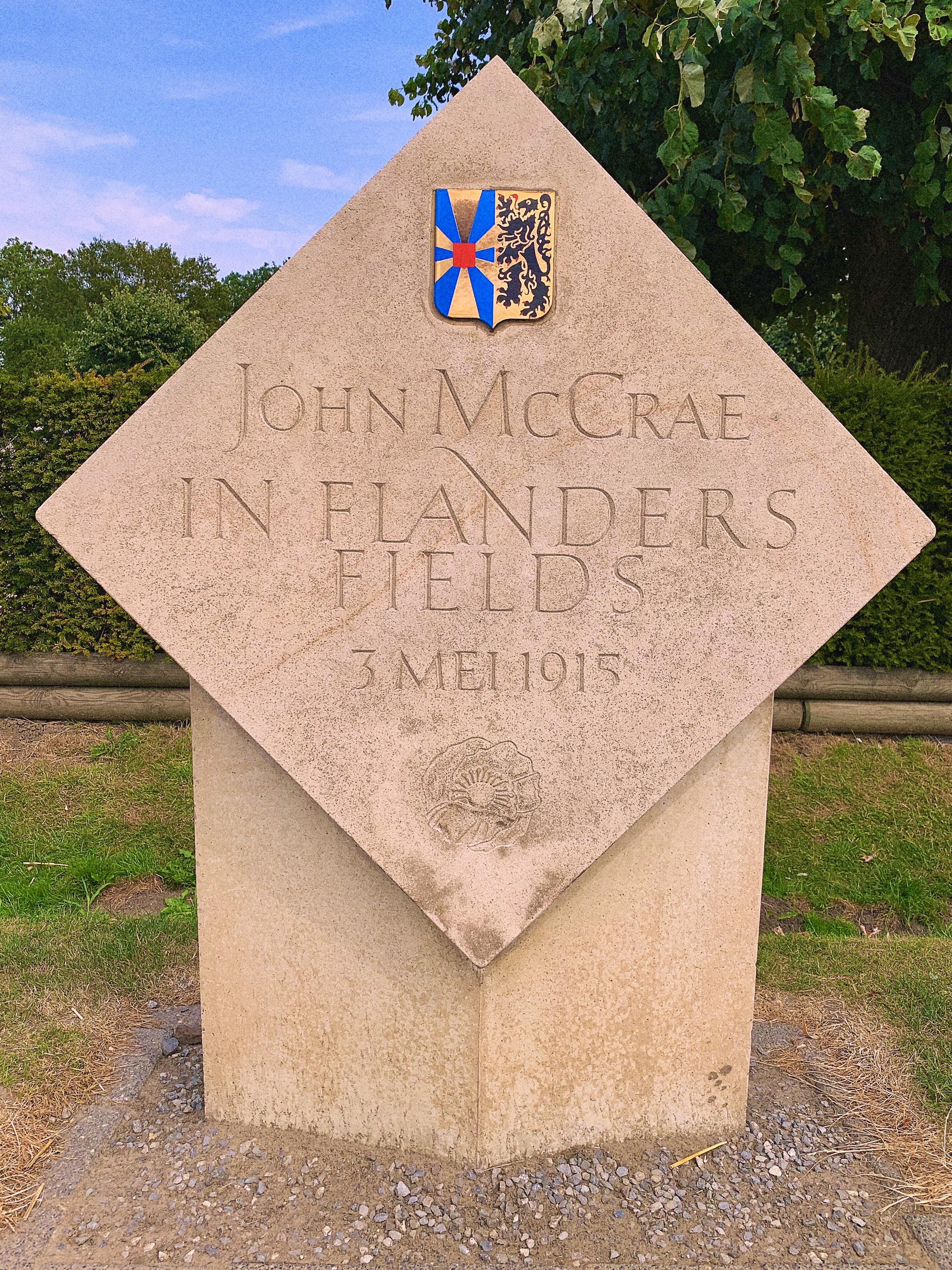 Stone monument with inscribed text "John McCrae IN FLANDERS FIELDS 3 MEI 1915" featuring an embossed badge on the top left depicting a blue windmill on a split background of blue and white with a gold design, positioned against a backdrop of green trees.