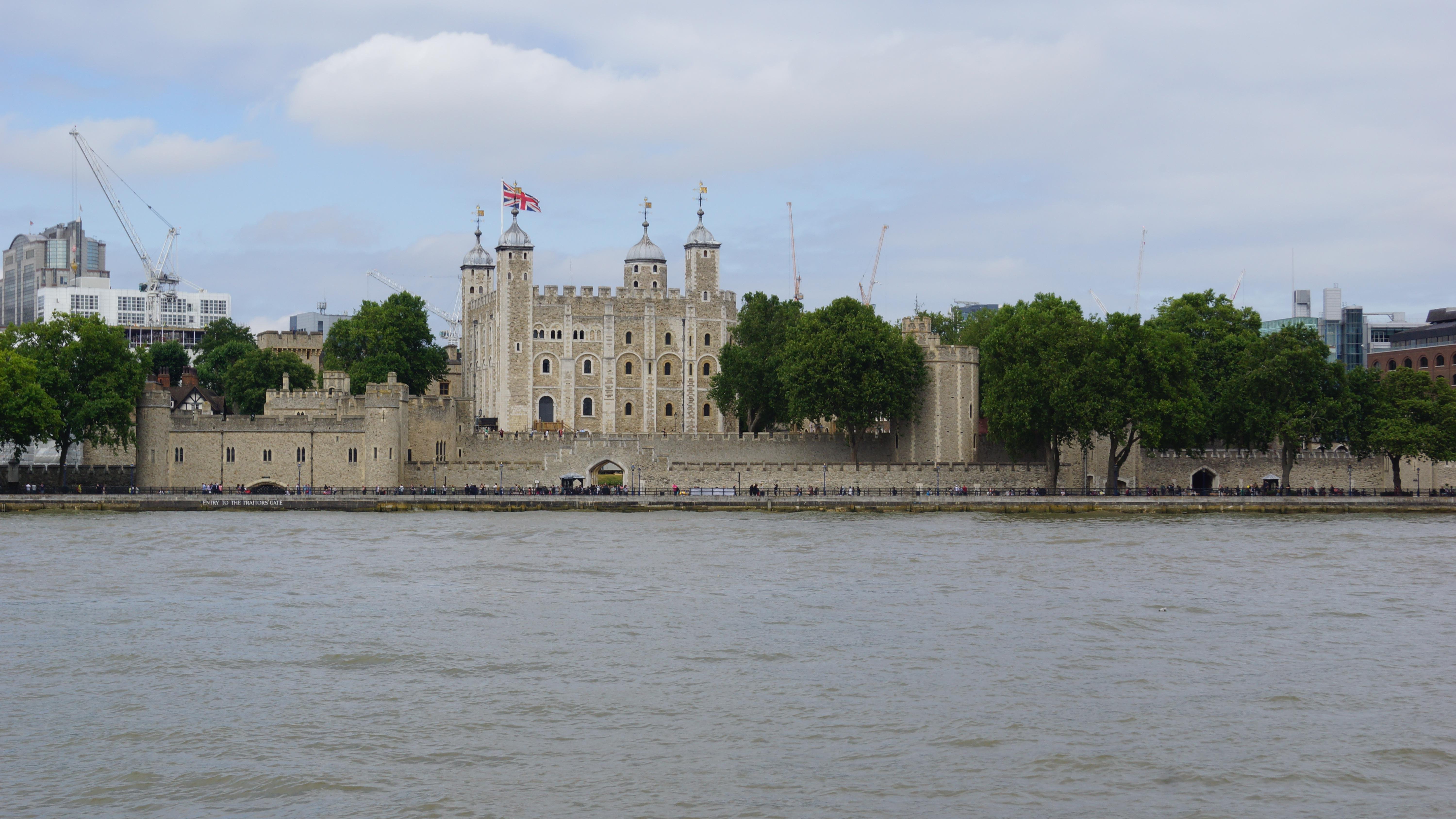 Wide view of the Tower of London as seen from across the River Thames, featuring its historic stone buildings and battlements under a cloudy sky. Several flags fly atop the central white tower.