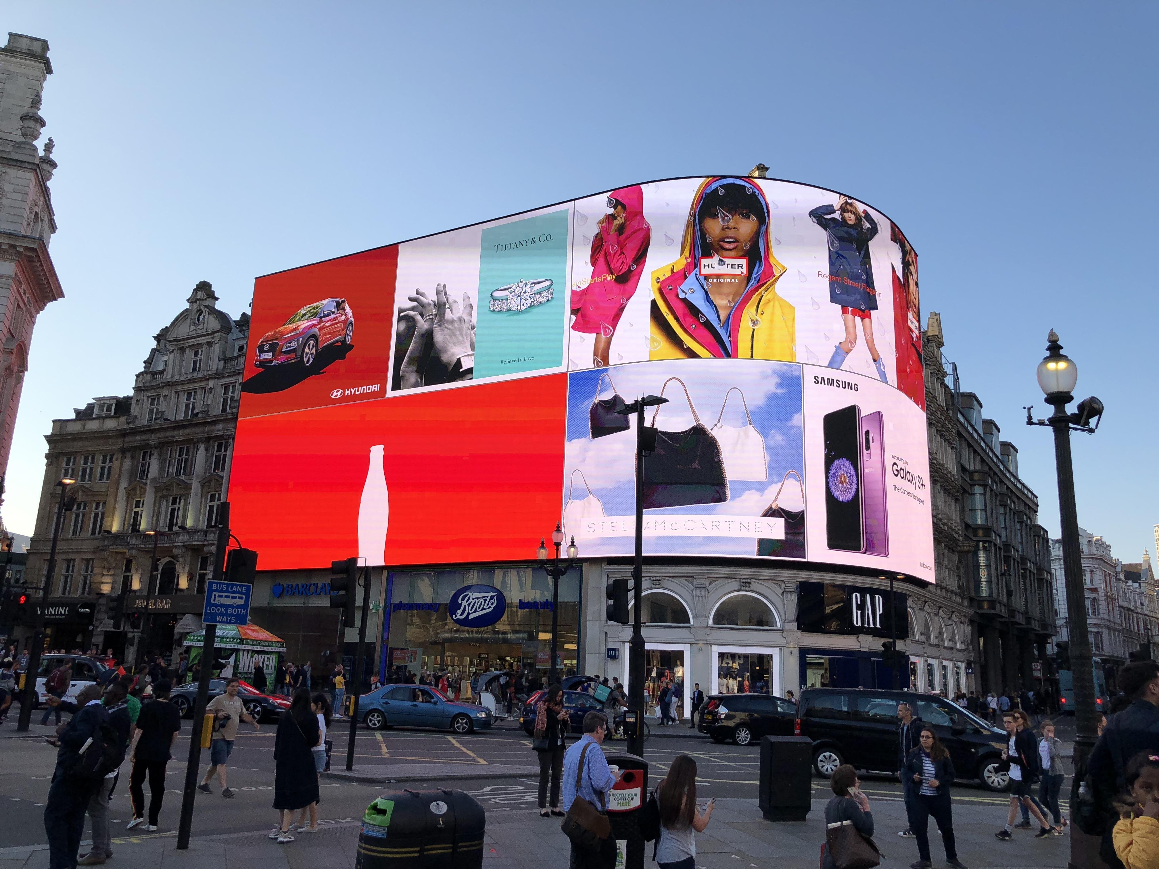 Large, curved electronic billboard displaying multiple advertisements dominates a busy city intersection during daytime. Ads include images of a car, fashion models, and electronic devices. Crowds of pedestrians and traffic are visible, with historic buildings under a clear blue sky in the background.