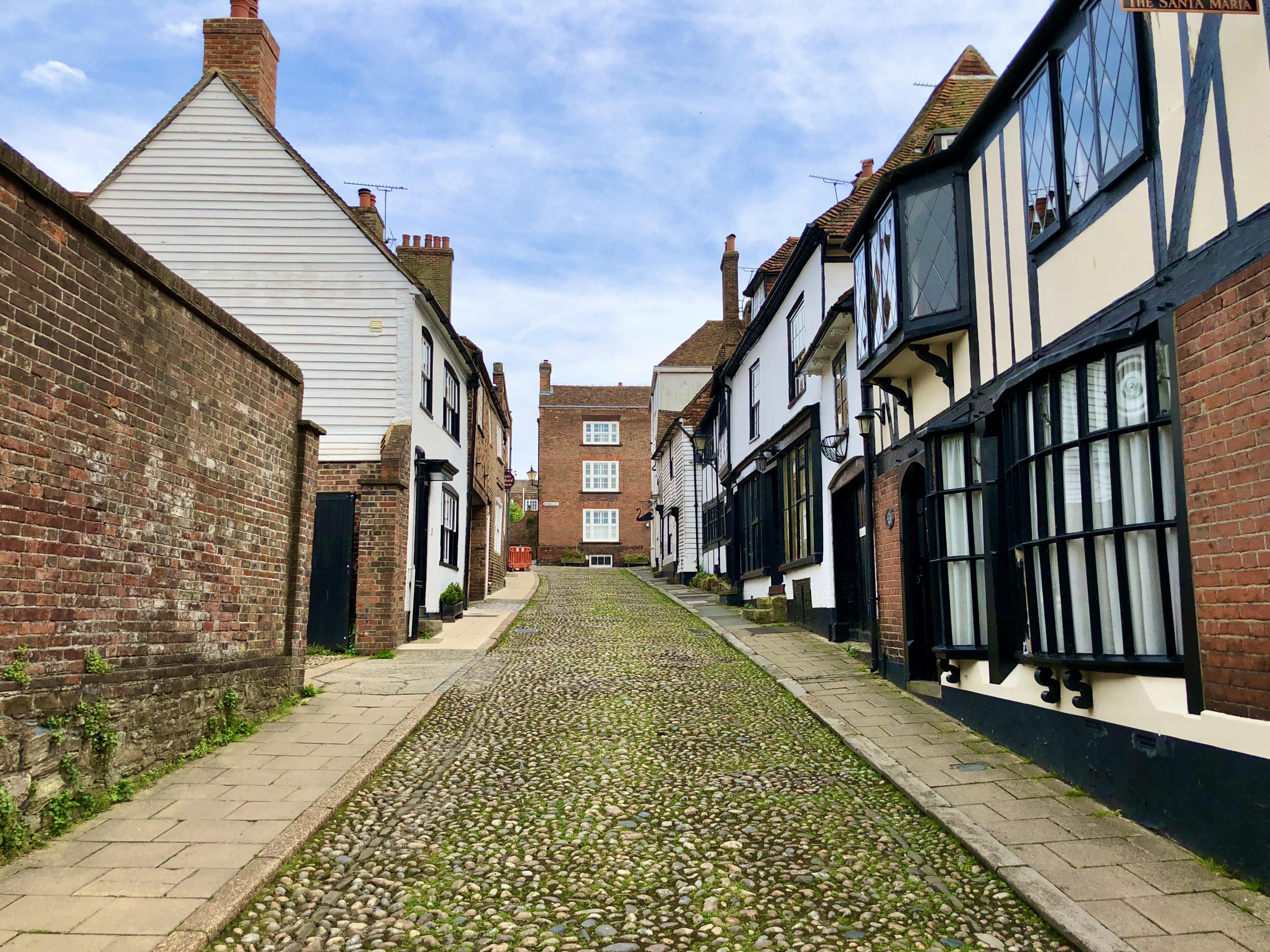 Cobblestone street flanked by traditional British houses with varying designs, including timber-framed and brick structures, under a clear sky. The street narrows towards the center where a red brick building stands prominently.