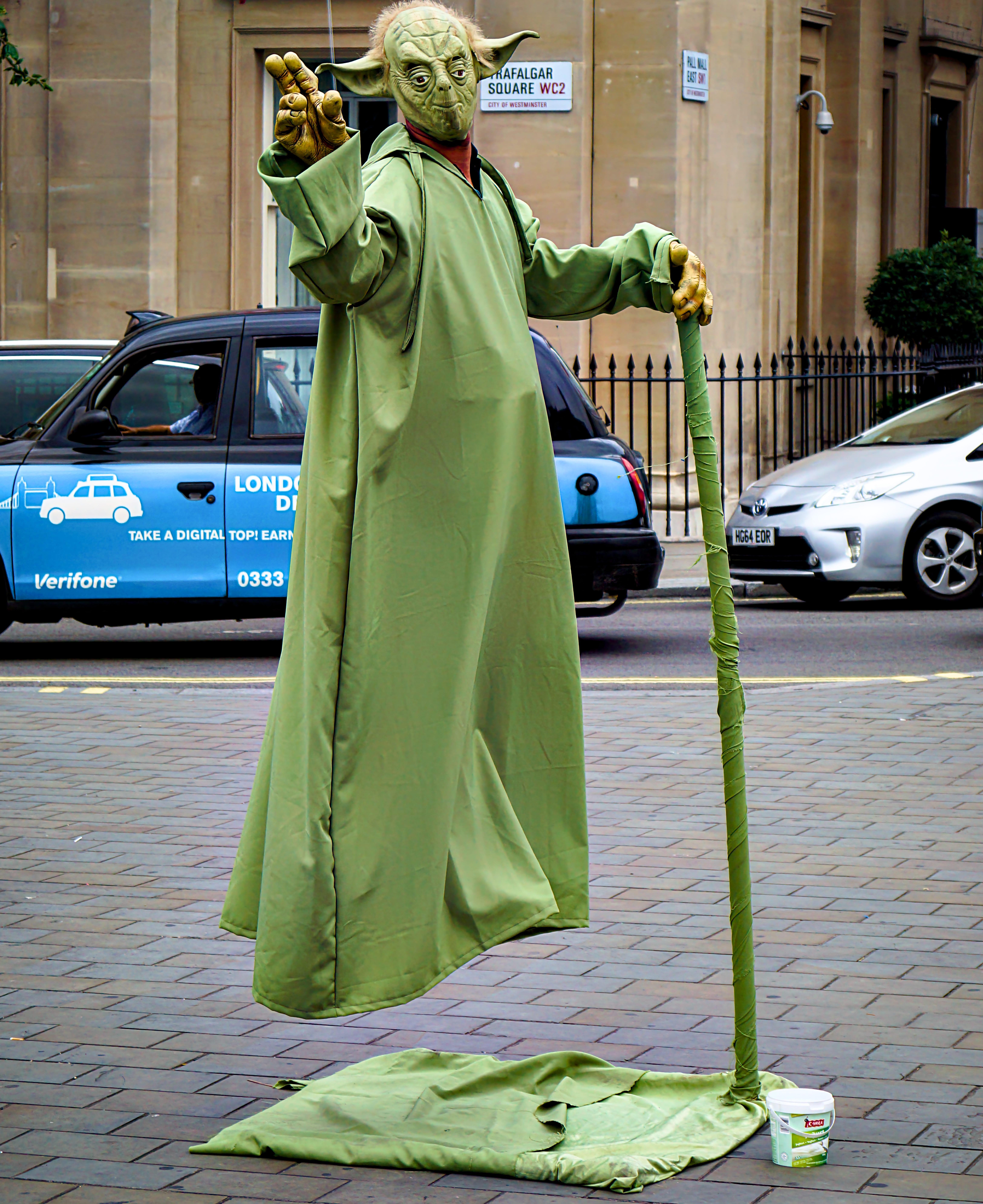 A person dressed as Yoda from Star Wars performs a levitation illusion on a city street. The performer wears a full Yoda mask and green robe, appearing to float above the ground with support from a green staff, which is wrapped in fabric matching the robe. The base of the staff touches the ground where a green fabric platform and a tip jar are placed. A London taxi and other cars are visible in the background, indicating an urban setting.
