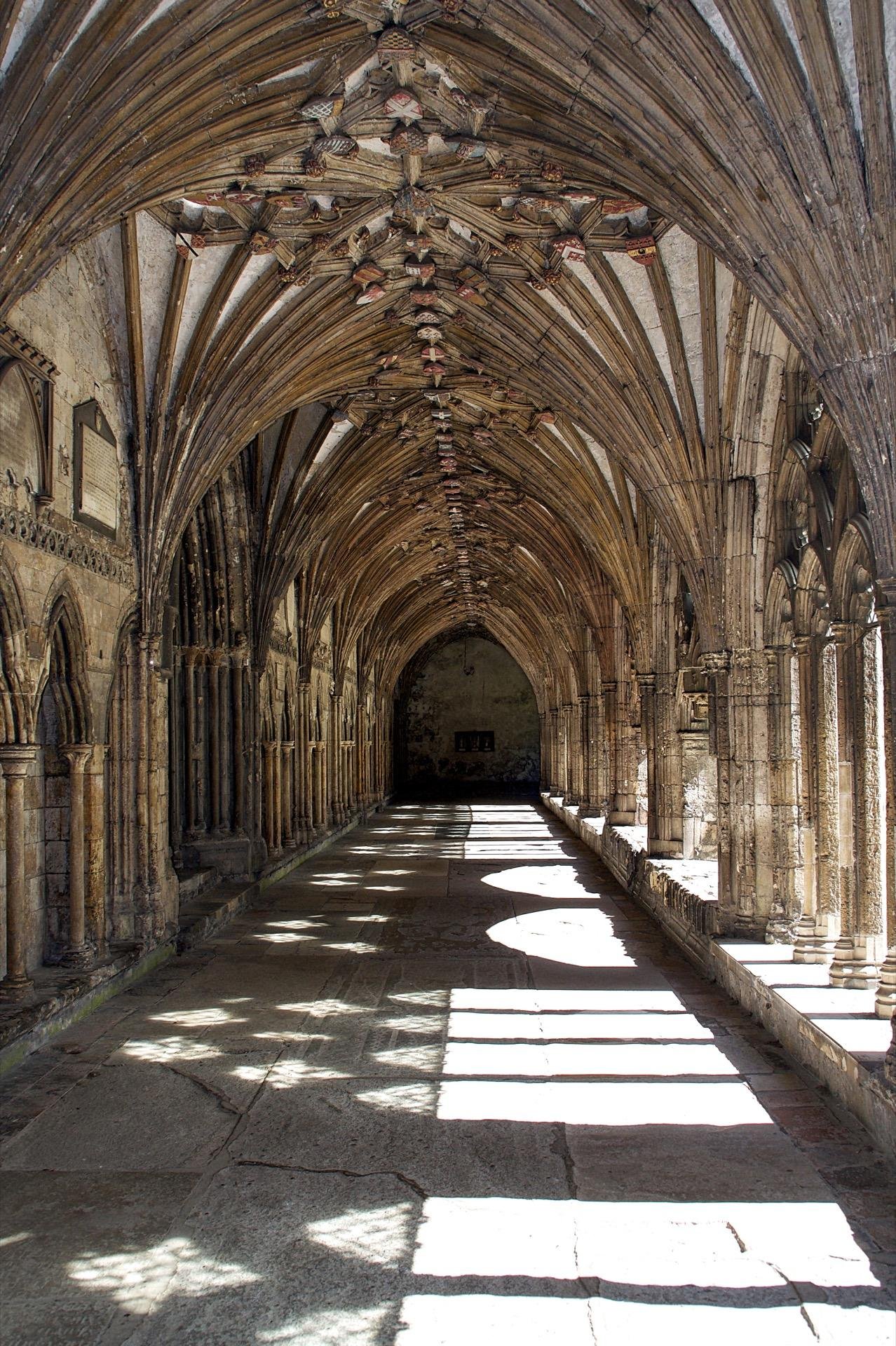 Gothic cloister with ornate, arched vault ceiling supported by columns. The weathered stone walls display carved arches and decorative elements. Sunlight filters through the arches, casting patterned shadows on the stone floor. At the far end, the passage appears darker with an obscured alcove.