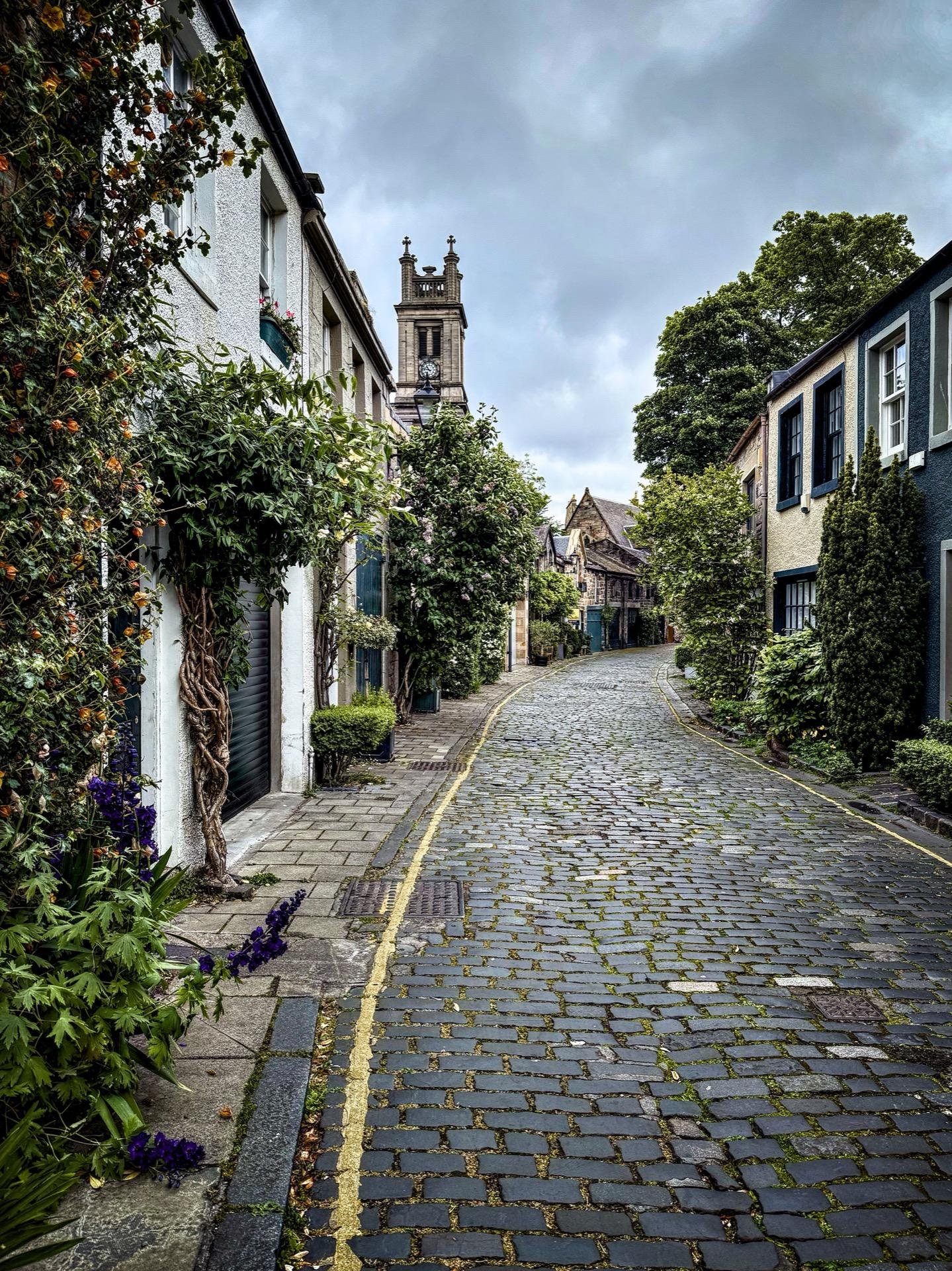Narrow cobbled street flanked by two-story houses, overcast sky, lush greenery on sides, flowers at curb, leading to a tower with a clock visible in the distance.