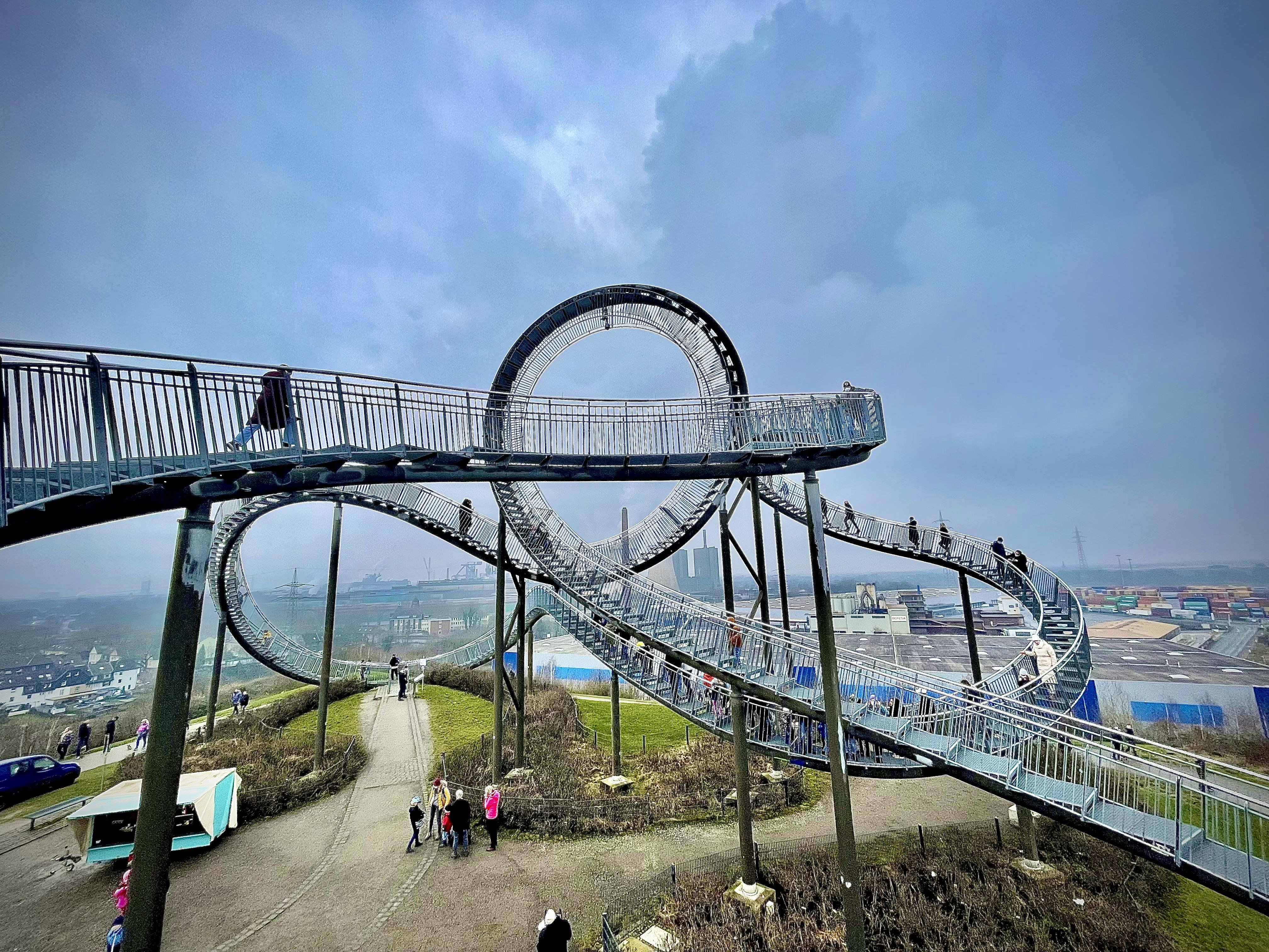 Sculptural walkway with complex loops, twists, and a circular structure, elevated above a grassy hill. People walk along the pathway, overlooking industrial buildings in the background under a cloudy sky.