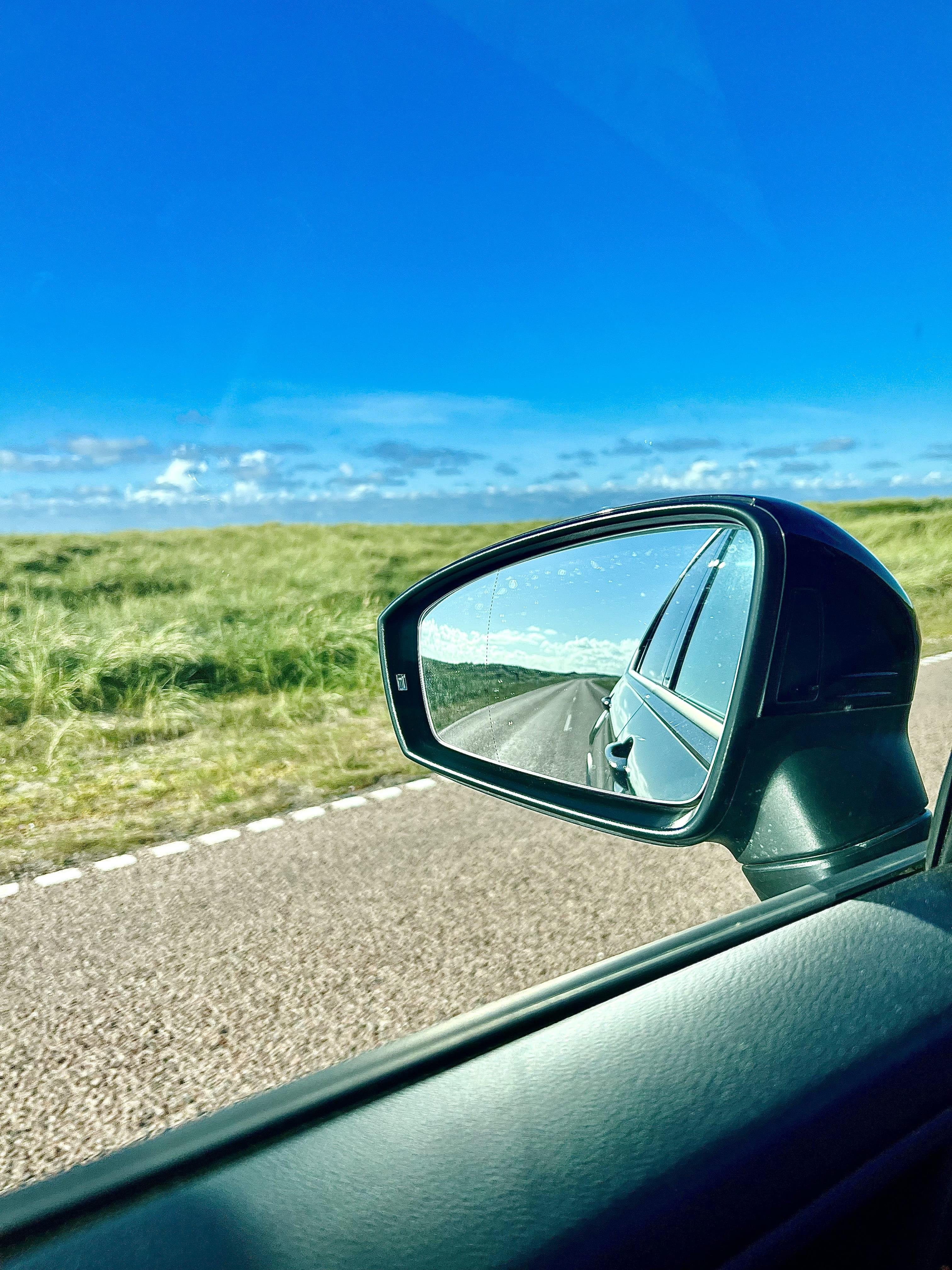 Car side mirror view of a road stretching back into the distance, reflecting clear sky and grassy fields beside the asphalt road. The foreground features the car's dark interior and part of the door.
