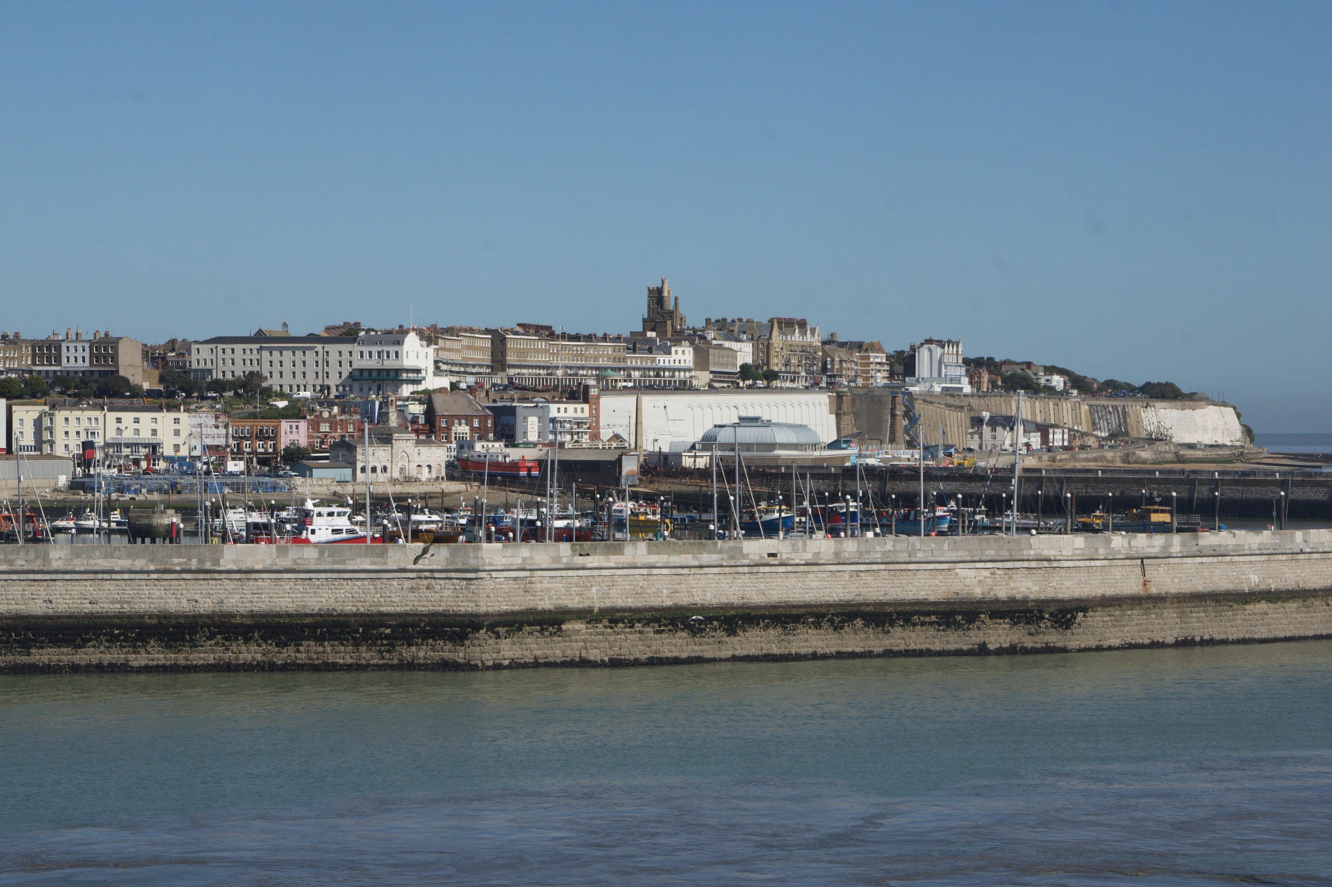 Photograph of a coastal town featuring a multi-tiered landscape with colorful buildings and historical structures on white cliffs. In the foreground, a harbor with several boats and cars is visible, separated from the town by a long seawall. Clear sky and calm blue water contribute to a serene seaside scene.