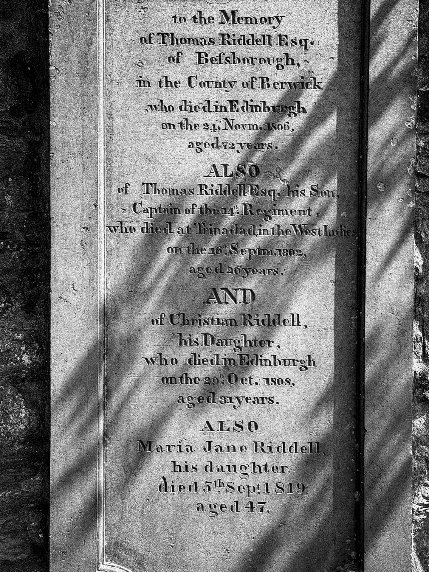 Vertical close-up of an aged stone memorial plaque with weathered, engraved text commemorating Thomas Riddell Esq. of Eelsborough and his family members, including dates of death and ages. The text is partially illuminated by sunlight casting shadows of leaves on the right side, highlighting the texture and erosion of the stone surface.