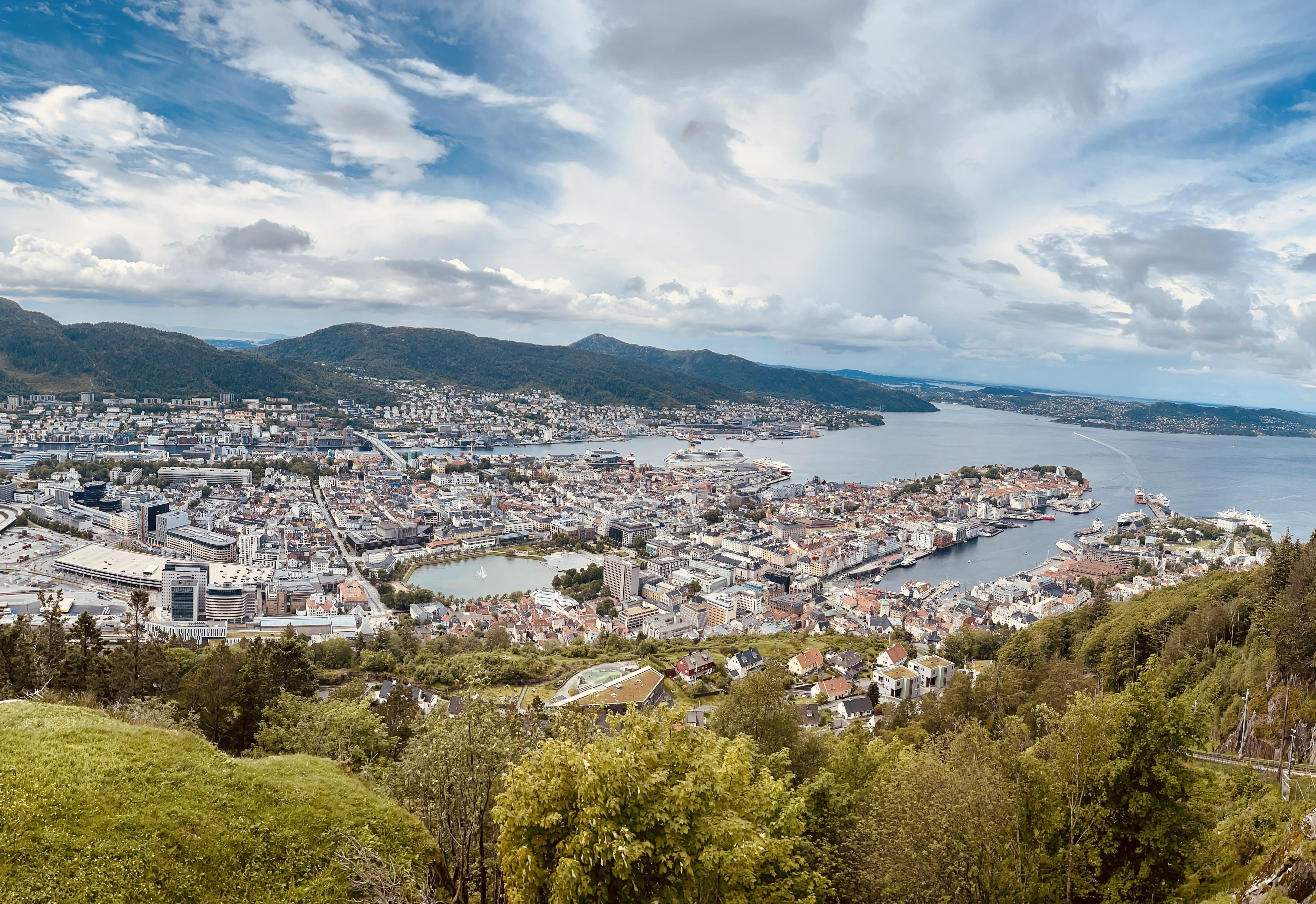 Panoramic view of a dense cityscape bordered by mountains and a wide river, featuring tightly packed buildings of various sizes, bridges over the water, and lush green areas at the foreground. The sky is partly cloudy over a serene natural and urban scene.