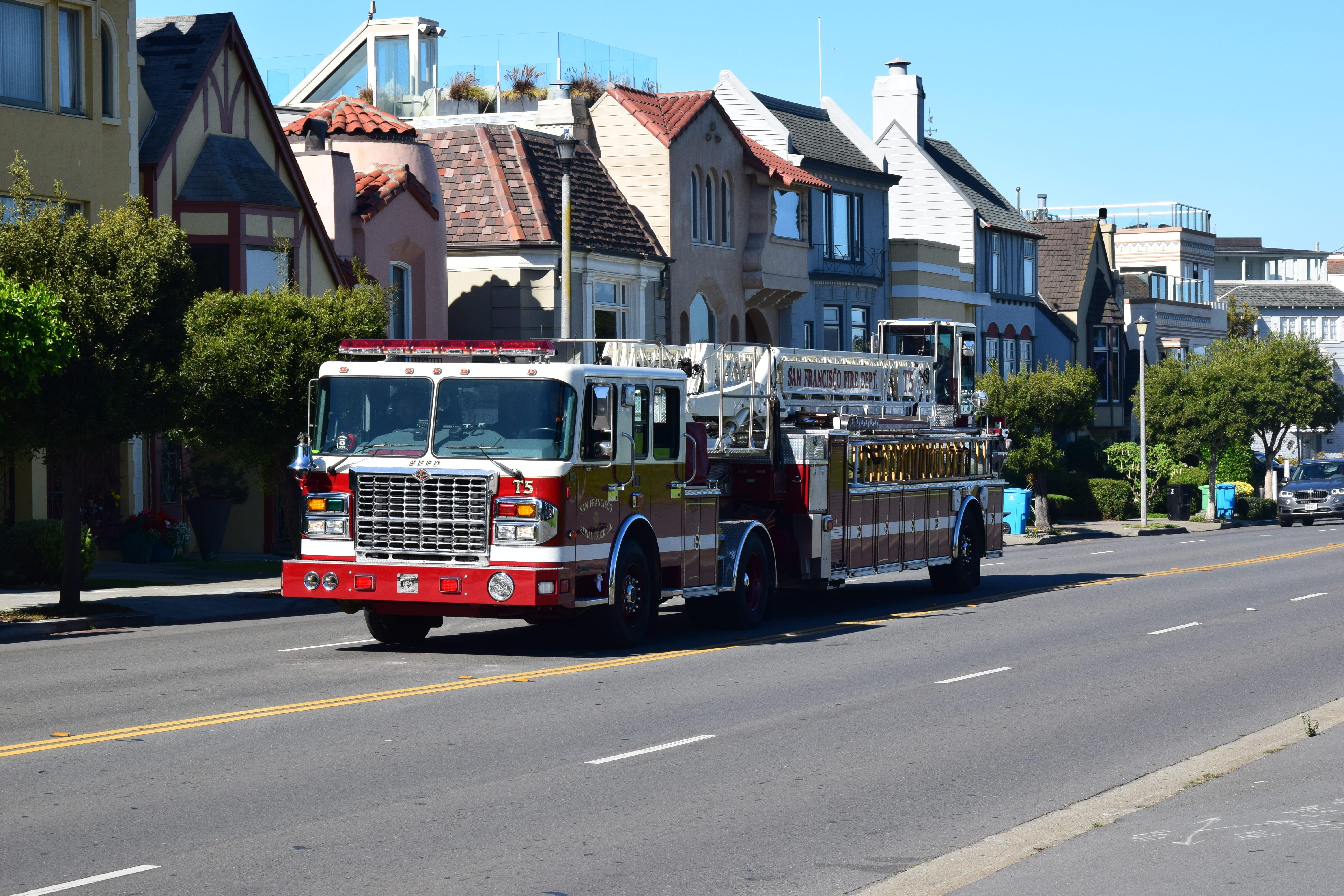 Red fire truck driving on a street flanked by two-story houses with varied architectural designs under a clear blue sky. The truck is marked with "15" and "San Francisco Fire Dept."