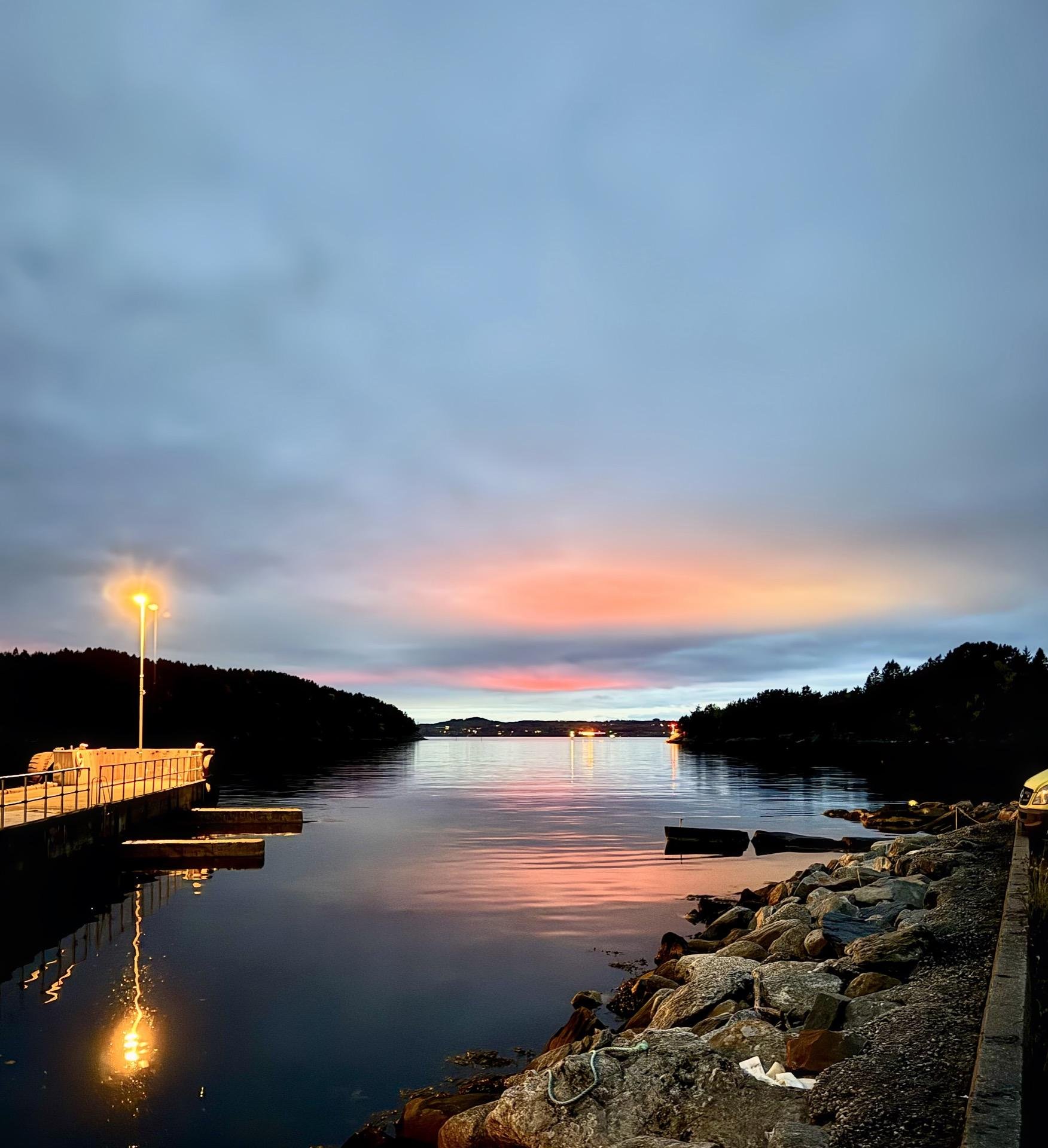 Twilight scene at a calm harbor with a lit lamp post on a pier to the left. Colorful sunset sky reflects on the water, which is bounded by a rocky shore in the foreground. Distant hills outline the horizon under the glowing sky.