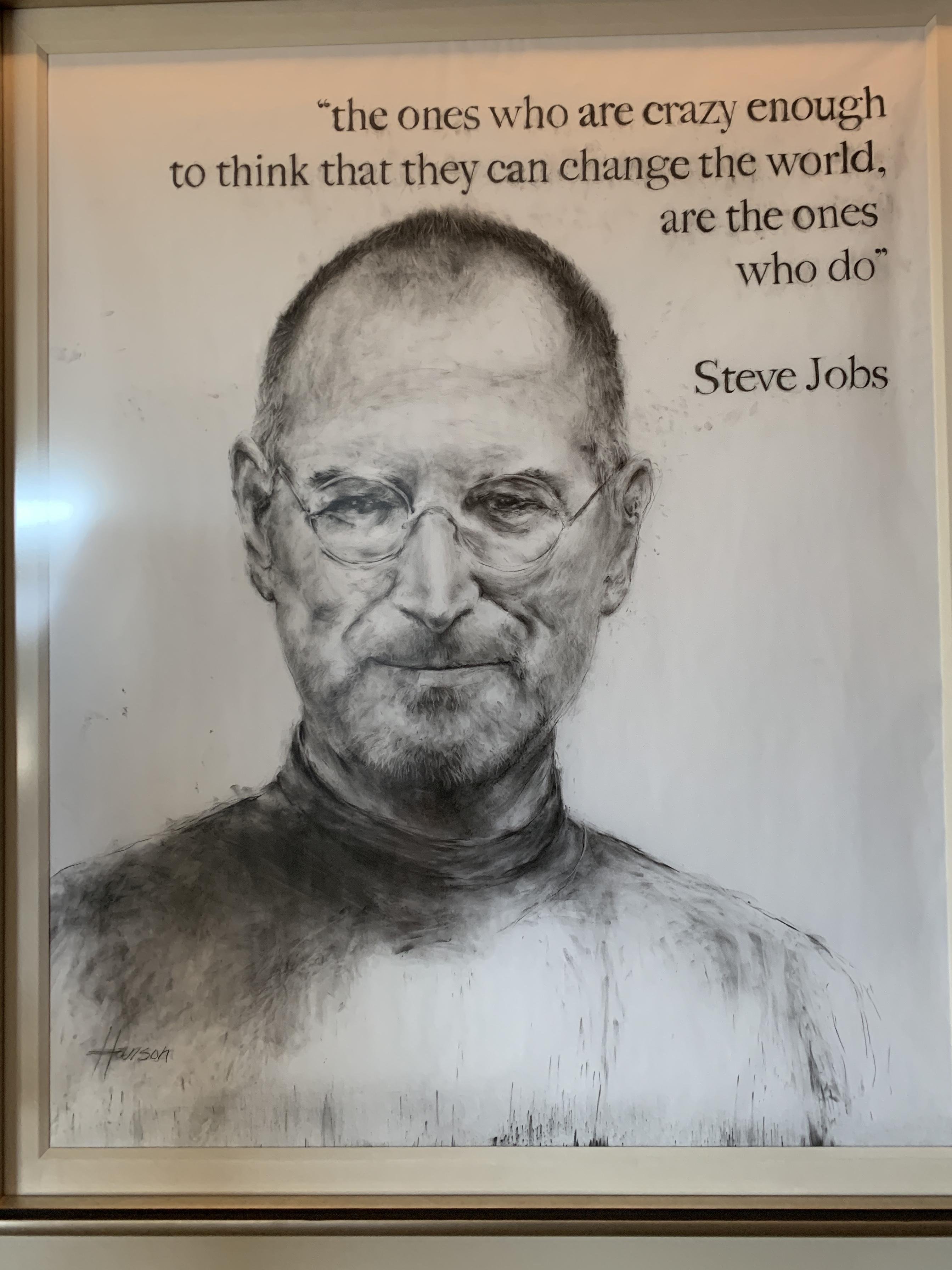 Black and white portrait of a man with glasses and closely cropped hair, wearing a turtleneck. Above the portrait, a quote reads: "the ones who are crazy enough to think that they can change the world, are the ones who do" attributed to Steve Jobs. The artist's signature appears at the bottom right of the drawing. The artwork is framed and displayed on a wall.